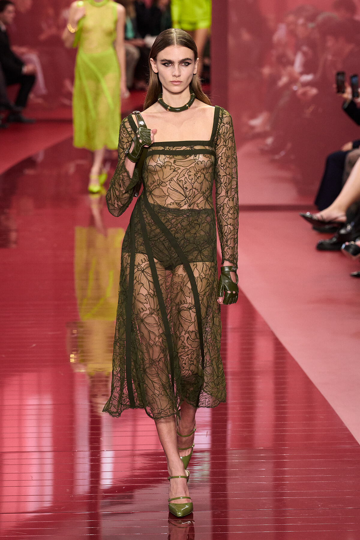Runway model in a sheer black lace dress with floral patterns, olive-green gloves, and a green choker walking a pink catwalk.