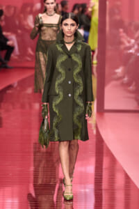 Model walks a pink runway in an olive-green coat with shimmering wavy fringe along the front, large lapels, and dark buttons, paired with matching gloves and handbag.