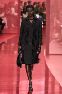 Model walking a black embellished coat down a pink runway, holding a fringed bag and wearing gloves.