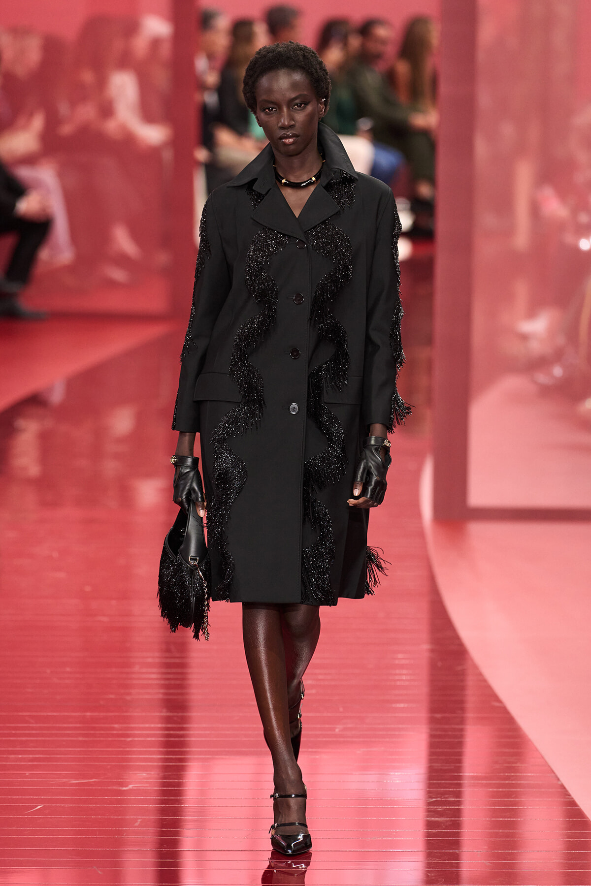 Model walking a black embellished coat down a pink runway, holding a fringed bag and wearing gloves.