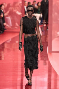 Model walks a glossy pink runway in a black fringed two-piece outfit with gloves and sunglasses.