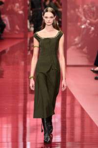 Model walking a runway in a dark olive green midi dress with front pockets, tie at the neckline, and black knee-high boots.