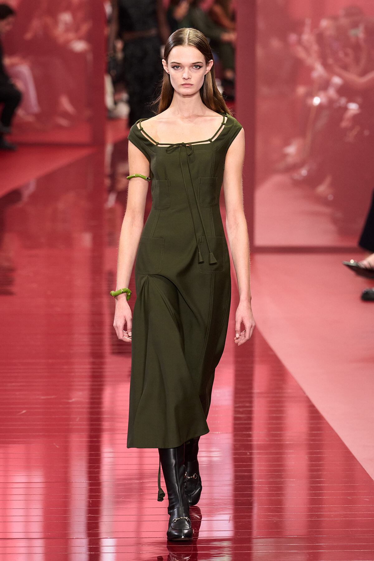 Model walking a runway in a dark olive green midi dress with front pockets, tie at the neckline, and black knee-high boots.