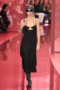 Model walks a pink runway wearing a black slip dress with a cutout, headscarf, sunglasses, knee-high boots, and a chain-strap bag, posed mid-stride.