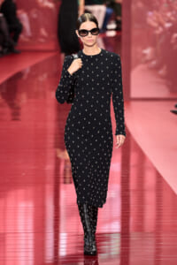 Model walking a red runway in a black patterned midi dress, oversized sunglasses, and knee-high studded boots, carrying a black bag.