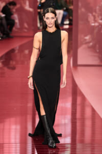 Model walks the runway in a sleeveless black gown with high neckline and dramatic side slits, paired with knee-high black boots and gold jewelry on a pink runway.