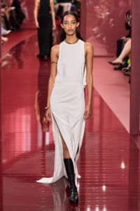 Model walking the runway in a white sleeveless gown with high neckline and high slits, paired with black boots and gold jewelry.