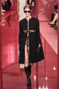 Model on a pink runway wearing a black long coat open at the waist, lace bodysuit, gloves, and oversized sunglasses.