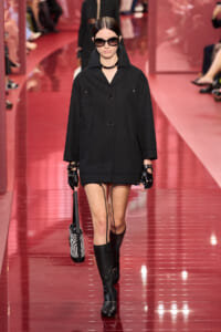 Model walking a runway in a black oversized coat, gloves, sunglasses, knee-high boots, and a zebra-striped handbag.