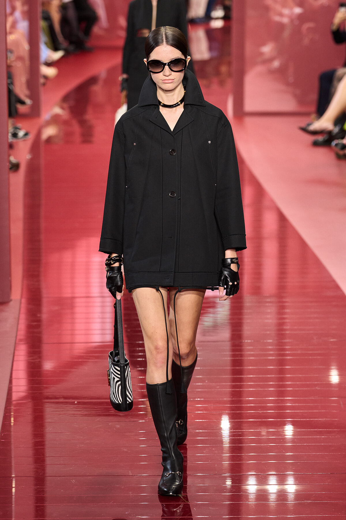 Model walking a runway in a black oversized coat, gloves, sunglasses, knee-high boots, and a zebra-striped handbag.