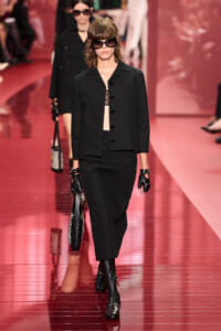 Model walks a fashion show in an all-black ensemble: blazer, midi skirt, over-the-knee boots, and large sunglasses, holding a striped bag.