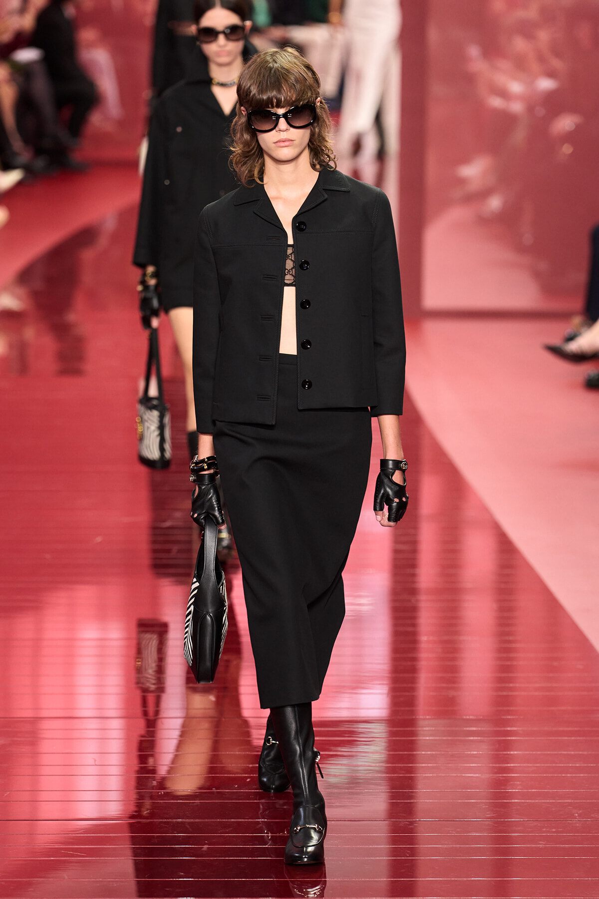 Model walks a fashion show in an all-black ensemble: blazer, midi skirt, over-the-knee boots, and large sunglasses, holding a striped bag.