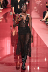 Model walks a sheer black lace dress on a pink/red runway, carrying a red bag and wearing knee-high boots.