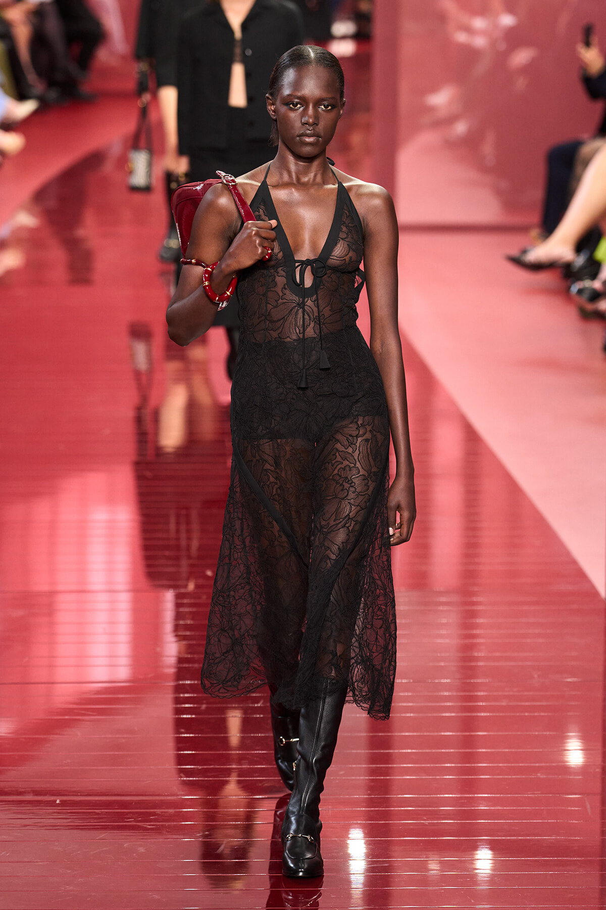 Model walks a sheer black lace dress on a pink/red runway, carrying a red bag and wearing knee-high boots.