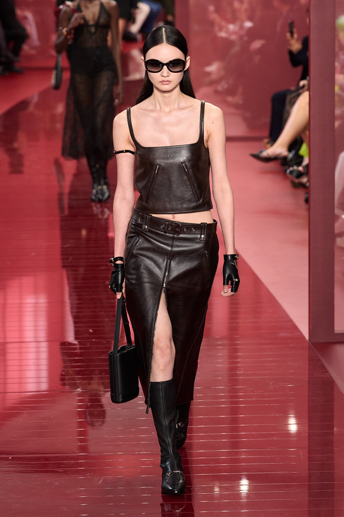 Model walking the runway in a black leather crop top and matching skirt, knee-high boots, gloves, and oversized sunglasses.