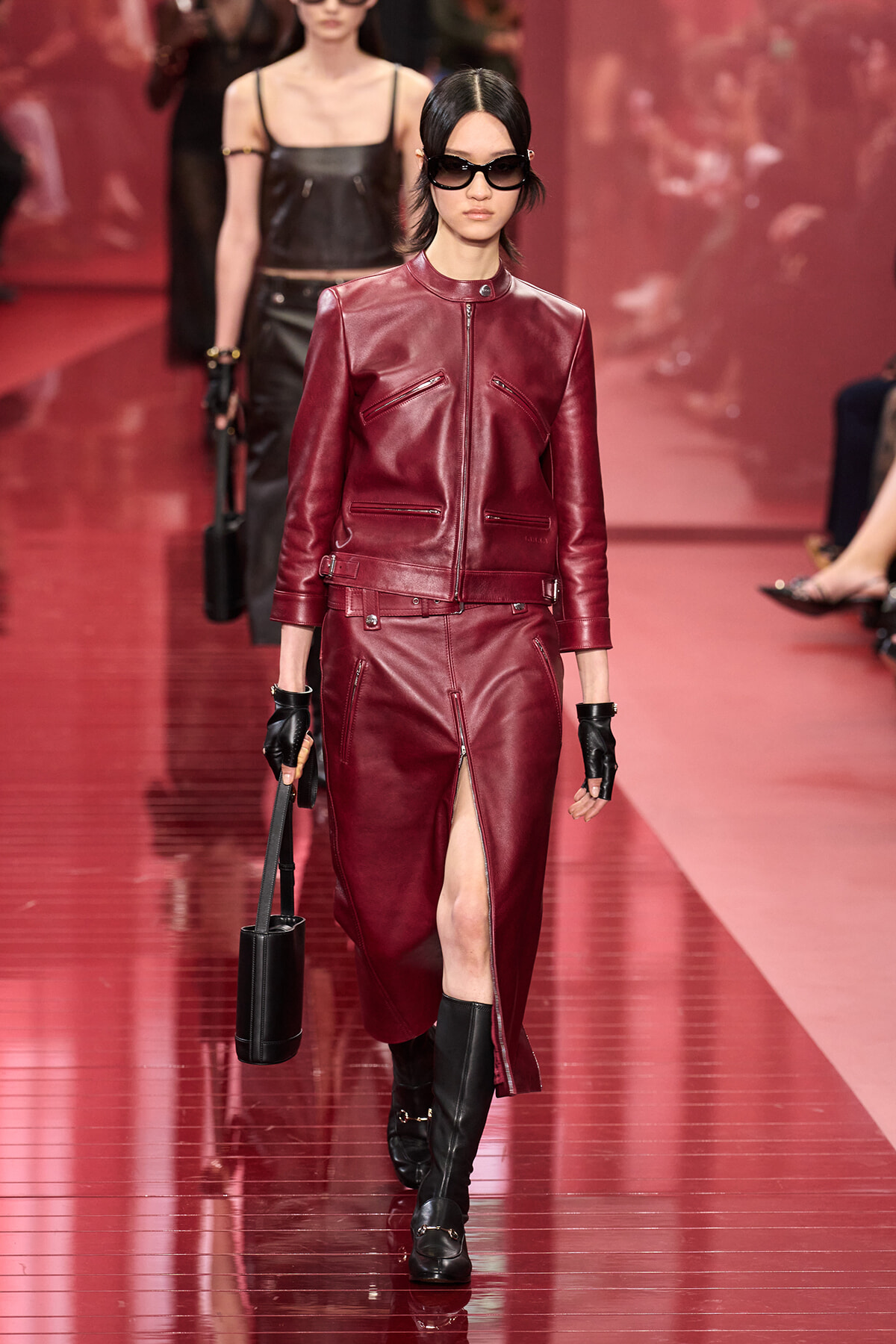 Model walking the runway in a burgundy leather jacket and matching midi skirt with a front slit, paired with black gloves, knee-high boots, and oversized sunglasses.