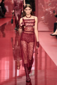 Model walking a red lace jumpsuit with sheer panels on a fashion runway, burgundy accessories and gloves visible