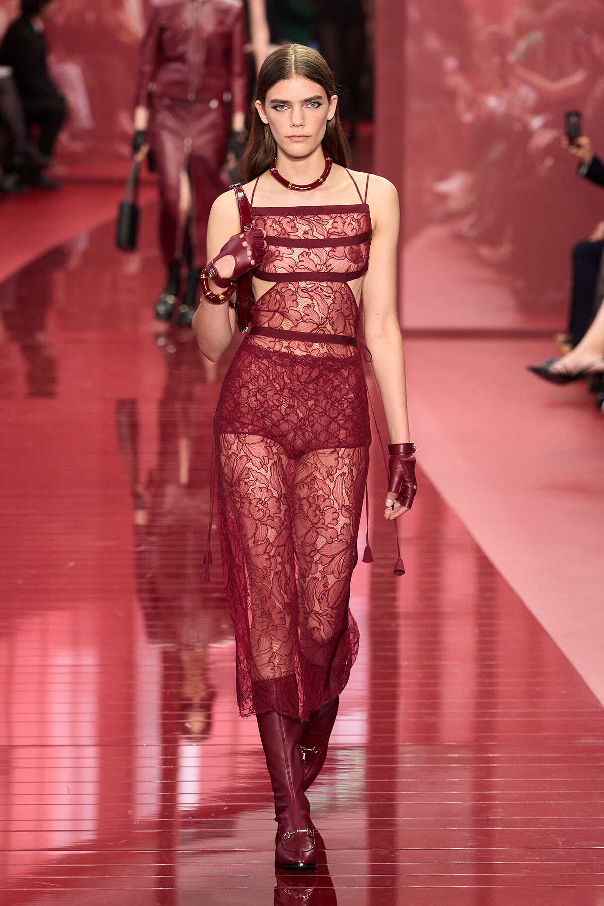 Model walking a red lace jumpsuit with sheer panels on a fashion runway, burgundy accessories and gloves visible