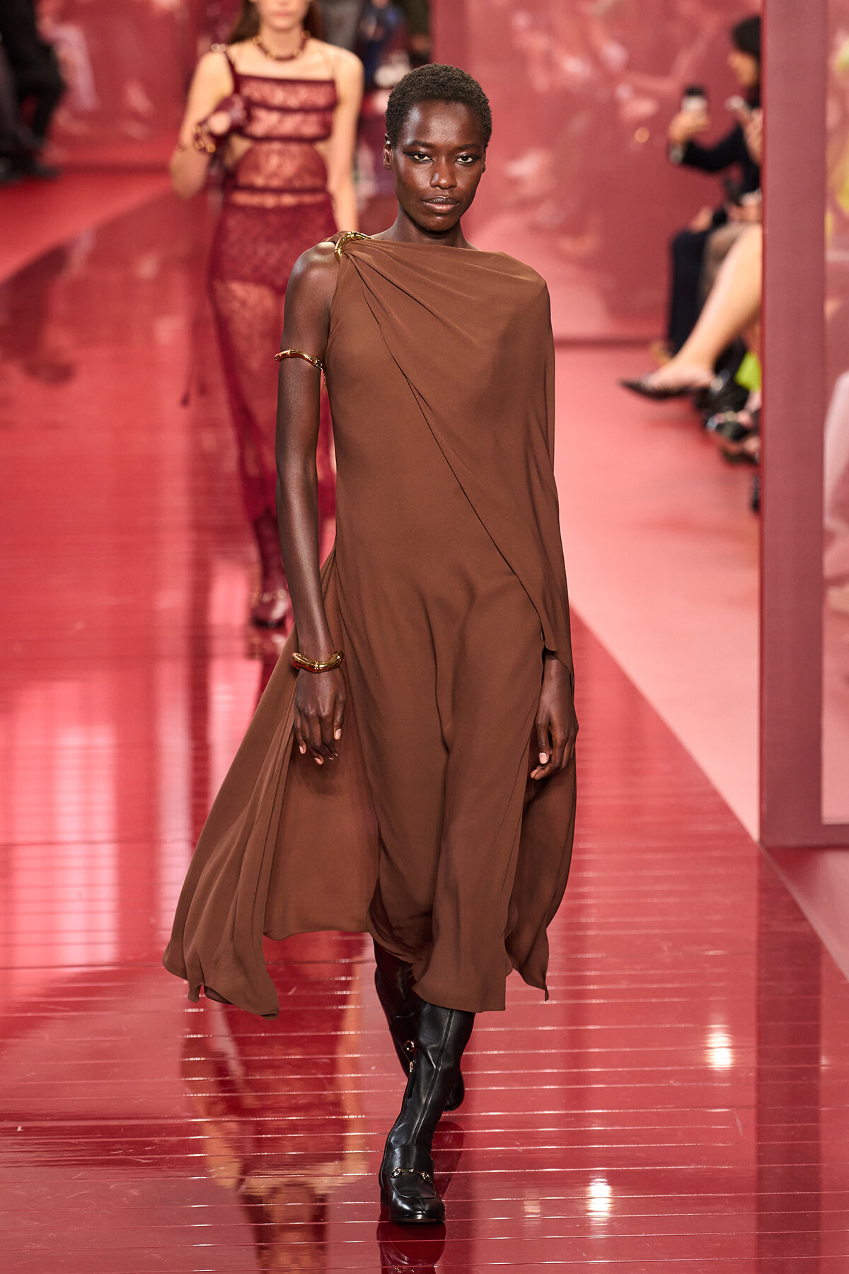 Model walking a pink glossy runway in a brown draped one-shoulder dress with gold bracelets and black boots.