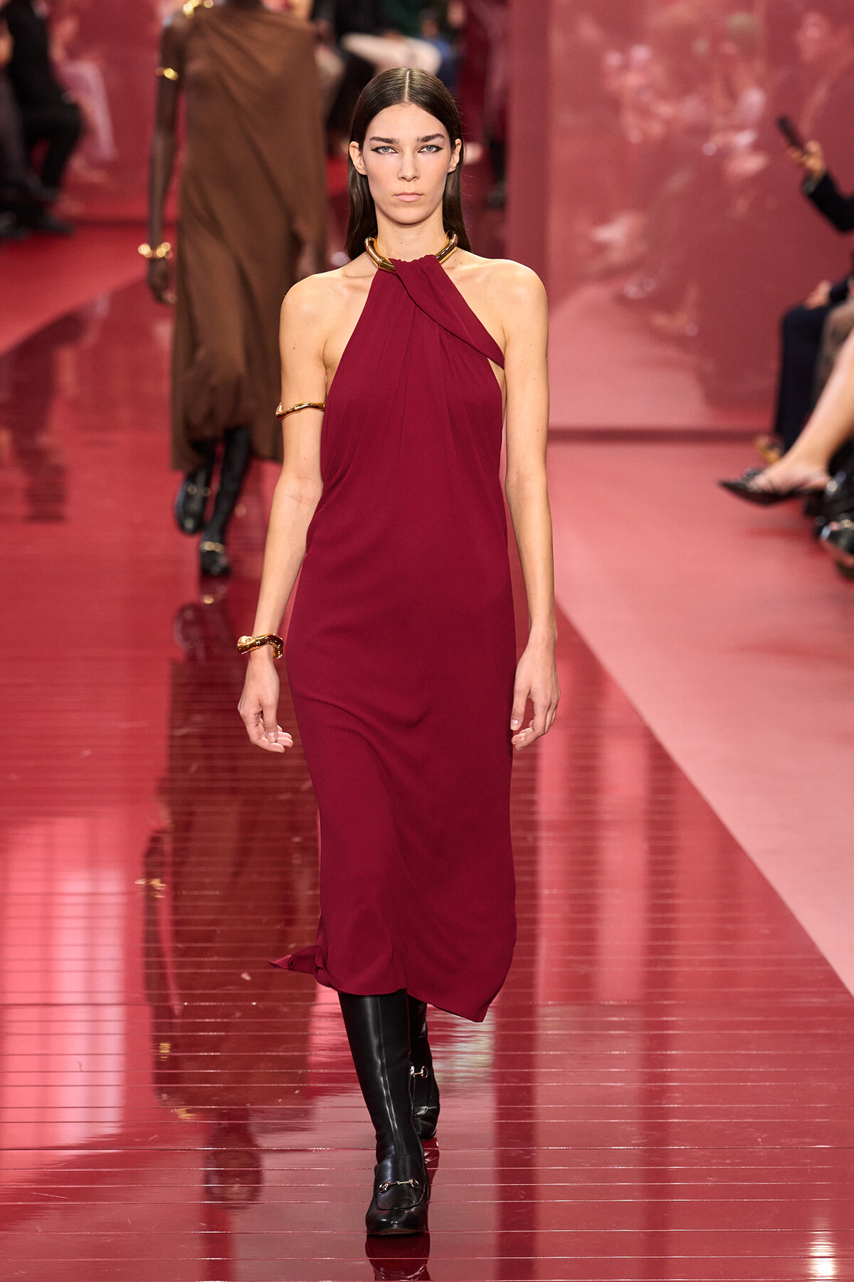 Model walks a burgundy halter dress with gold choker and bracelets on a reflective pink runway with other designs in the background.