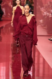 Model walks a burgundy fashion show wearing a matching oversized button-down shirt and wide trousers with a belt and bold necklace.