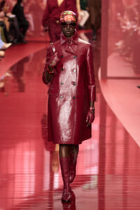 Model walking a fashion runway in a burgundy patent leather trench coat, matching gloves and boots, with a colorful headscarf and dark sunglasses.
