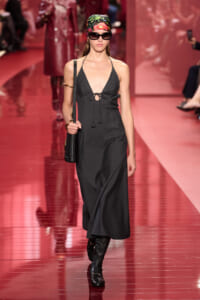 Model struts the runway in a black halter dress with a front tie, floral headscarf, sunglasses, and knee-high boots.