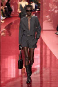 Runway model in a dark gray romper with a plunging neckline, black belt, knee-high boots, sunglasses, and gold jewelry walking a red glossy catwalk.