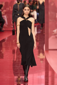 Model walking a runway in a black halter dress with a central cutout and gold accessories, knee-high boots.