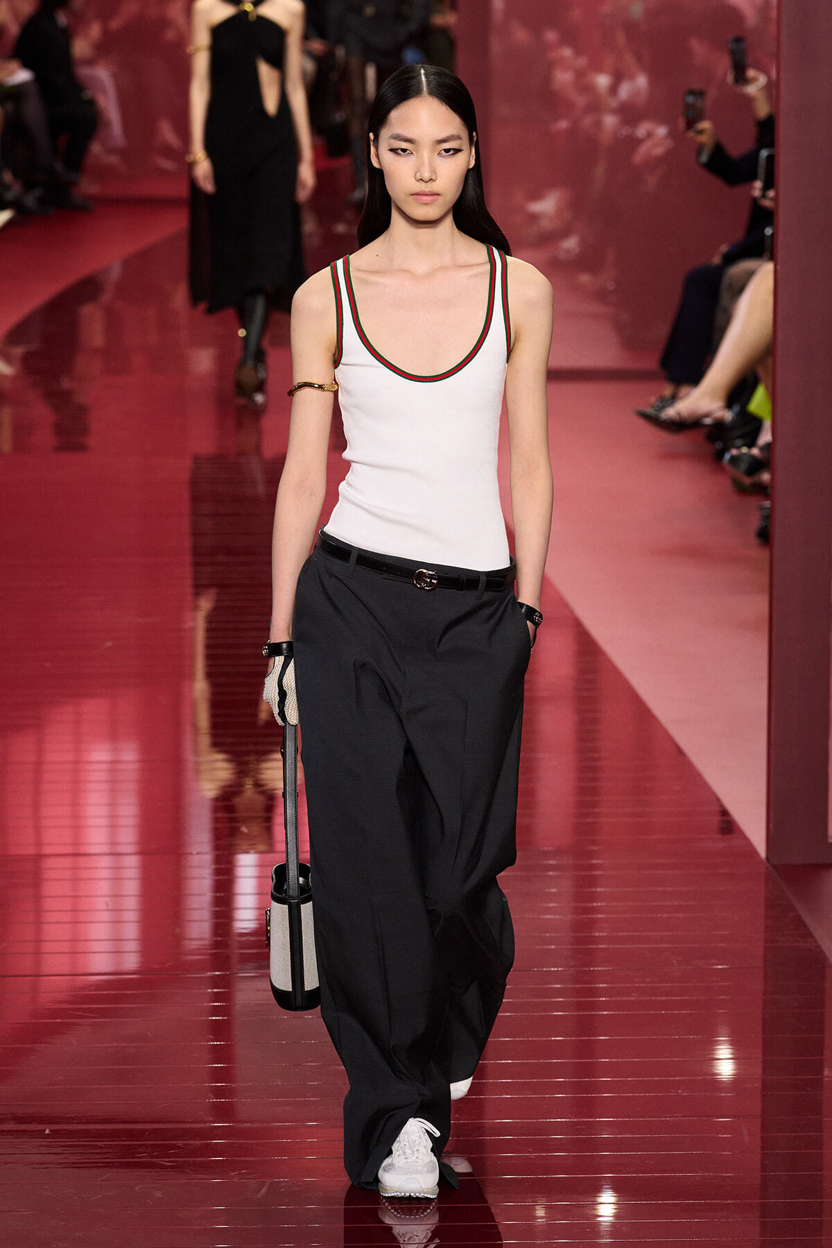 Model walking a fashion runway in a white racerback top with green-red trim and oversized black pants, carrying a small striped bag.