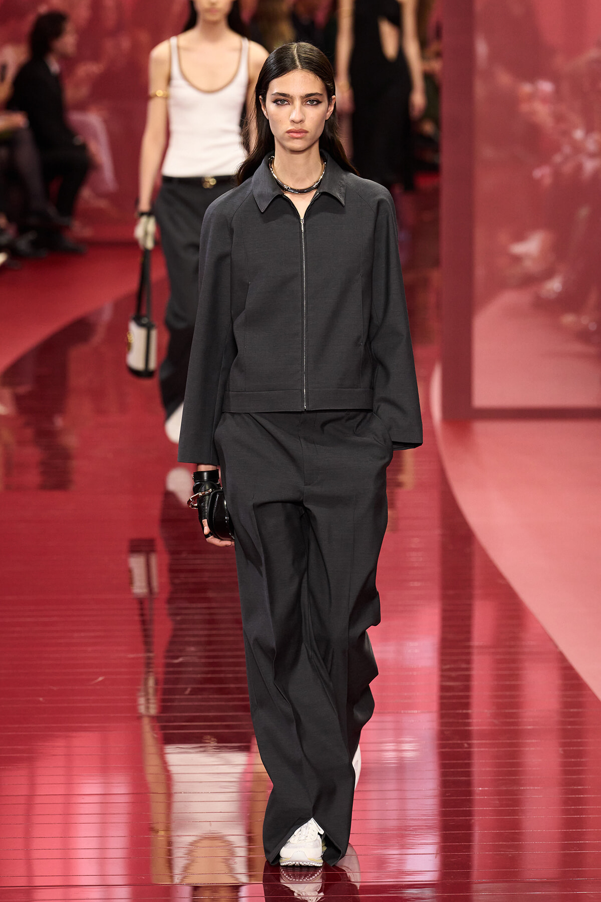 Model walking a glossy red runway in a dark gray zip-front jacket and matching wide-leg pants, white sneakers, with a choker necklace.
