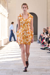 Model walking a runway in a bright orange floral print dress with a square neckline and pockets, during a fashion show in a sunlit venue.