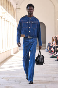 Male model walking a fashion runway in a blue denim jacket and jeans, carrying a black leather duffel bag.