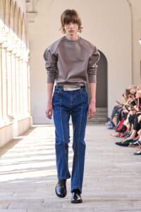 Model walking a runway in a gray oversized sweatshirt, high-waisted blue jeans, and a wide belt, black boots, in a sunlit colonnade.