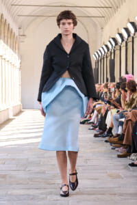 Model walking a fashion runway in a black blazer and light blue skirt in a stone-arched venue with seated audience to the side.