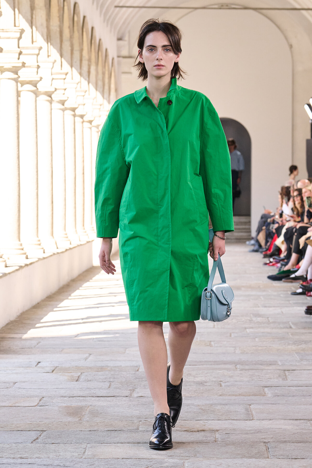 Model walks a runway in a bright green knee-length coat, with a light blue handbag and black shoes.