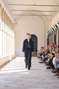 Model in a navy suit walks the runway through a sunlit arched corridor as spectators film with smartphones on the side.