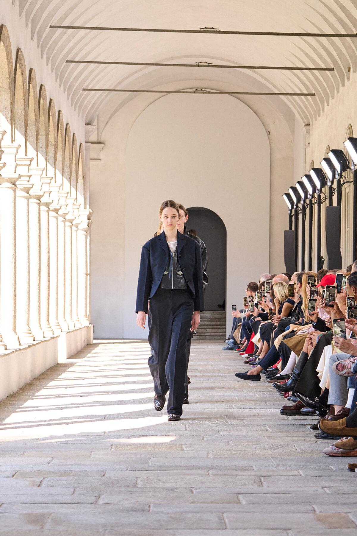 Model in a navy suit walks the runway through a sunlit arched corridor as spectators film with smartphones on the side.