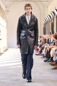 Male model walking a runway in a knee-length black leather coat over a white shirt and navy trousers; audience sits along both sides in a bright arcade venue.