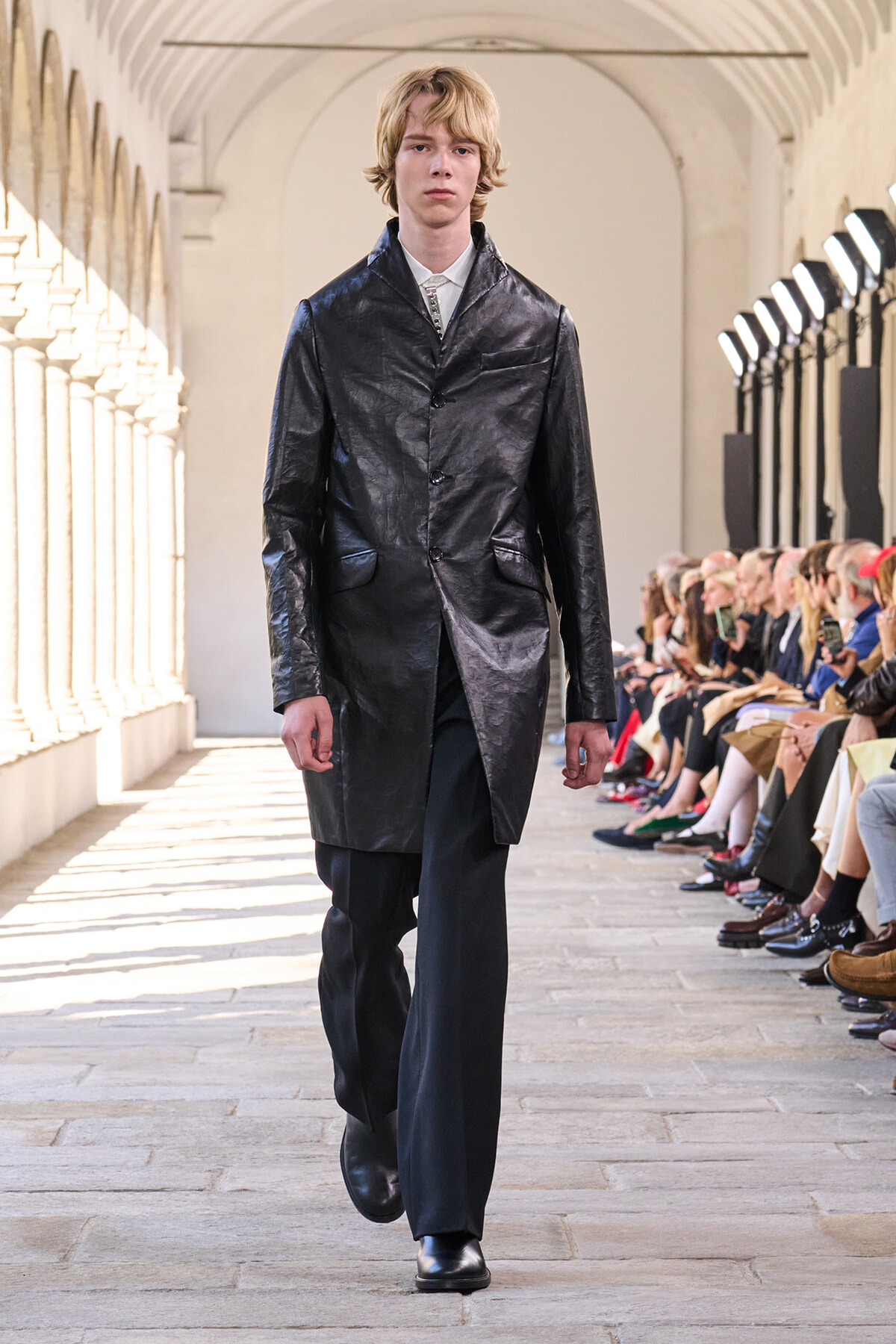 Male model walking a fashion runway in a shiny black leather trench coat, black pants, and boots, audience on both sides.