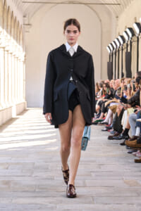 Model walking a runway in a black oversized blazer over a white shirt with black shorts and a light blue handbag. Audience visible on the sides of the catwalk.