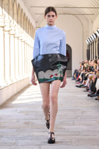 Model on a runway wearing a light blue high-neck blouse with a black, sculptural mini skirt featuring a painted landscape (trees, hills).