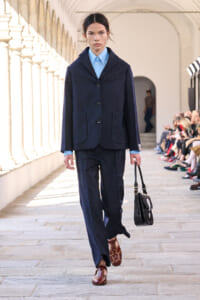 Female model walking a runway in a navy pinstripe suit with a light blue shirt, carrying a black handbag and wearing burgundy studded shoes.