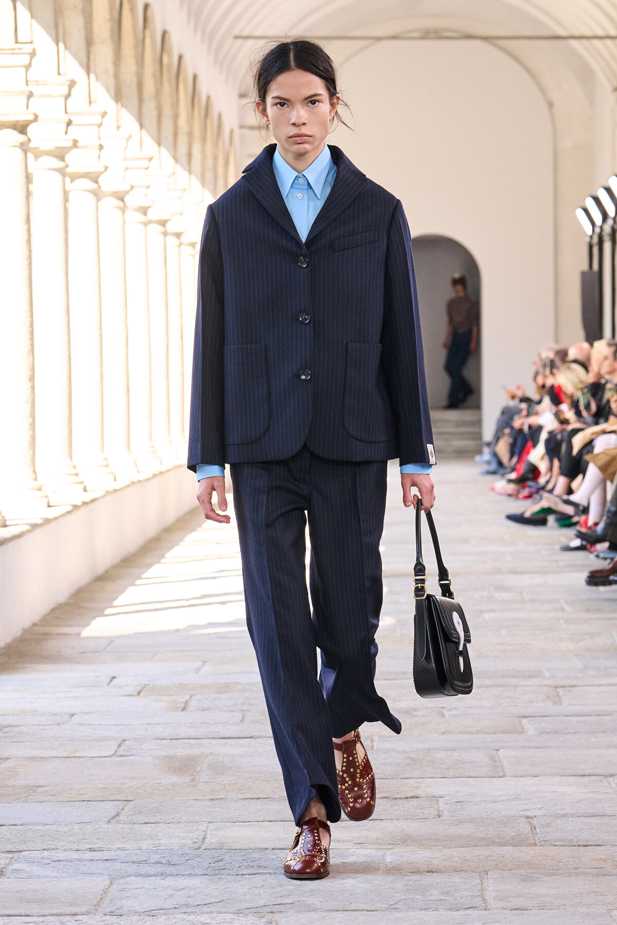 Female model walking a runway in a navy pinstripe suit with a light blue shirt, carrying a black handbag and wearing burgundy studded shoes.