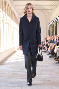 Model on a runway wearing a navy knee-length coat with buttons, gray trousers, and brown shoes, carrying a small black handbag with red trim as the audience watches fly by.