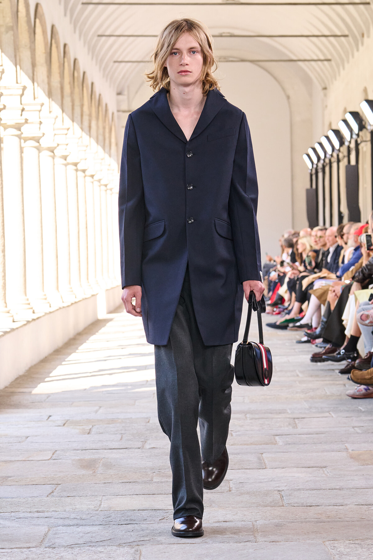 Model on a runway wearing a navy knee-length coat with buttons, gray trousers, and brown shoes, carrying a small black handbag with red trim as the audience watches fly by.