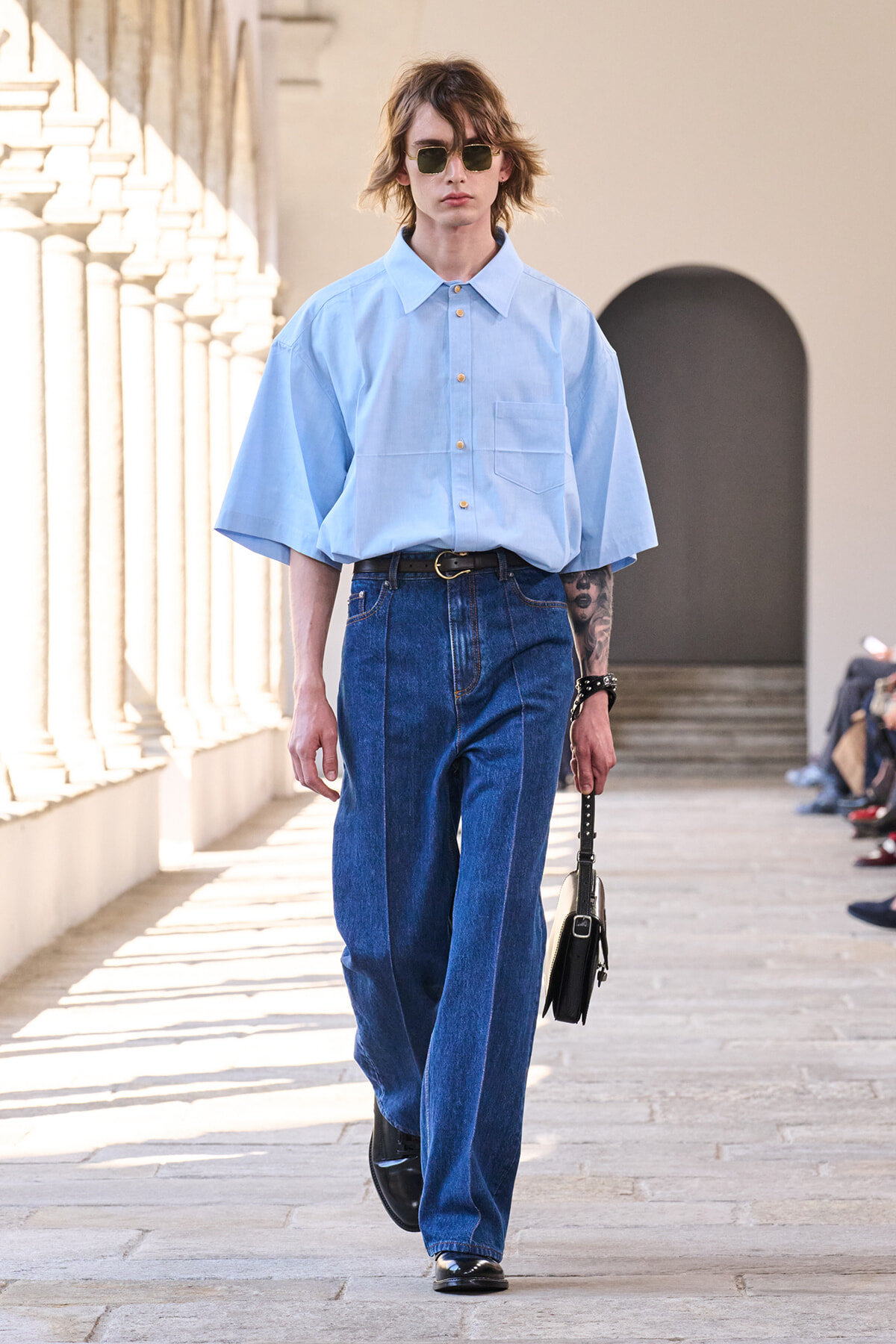Model walking a fashion show in a light blue oversized shirt, high-waisted wide-leg jeans, and a small black handbag.
