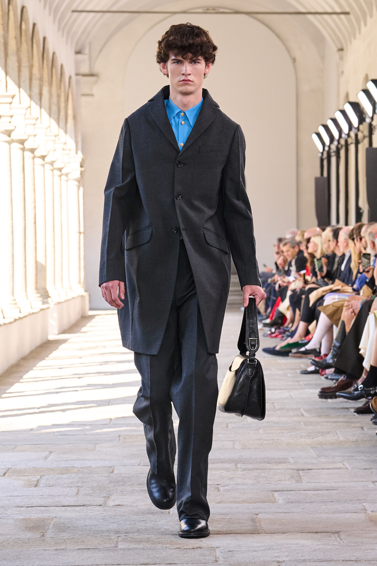 Male model walking a runway in a dark overcoat and matching trousers, bright blue shirt, and a black leather bag in hand.