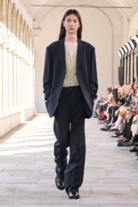 Model walks a runway in a dark pinstripe suit with an olive vest and mesh top, audience seated along the sides.
