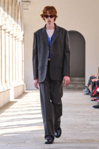 Male model walking a fashion runway in a oversized gray suit with blue inner top and dark sunglasses, in a bright architectural venue